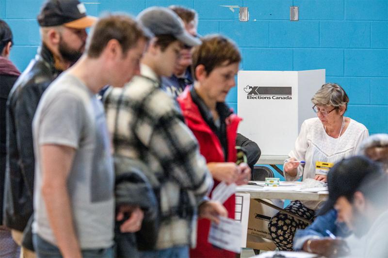 Several people line up at a polling station in a gymnasium as a volunteer sits in front of the cardboard polling booth.