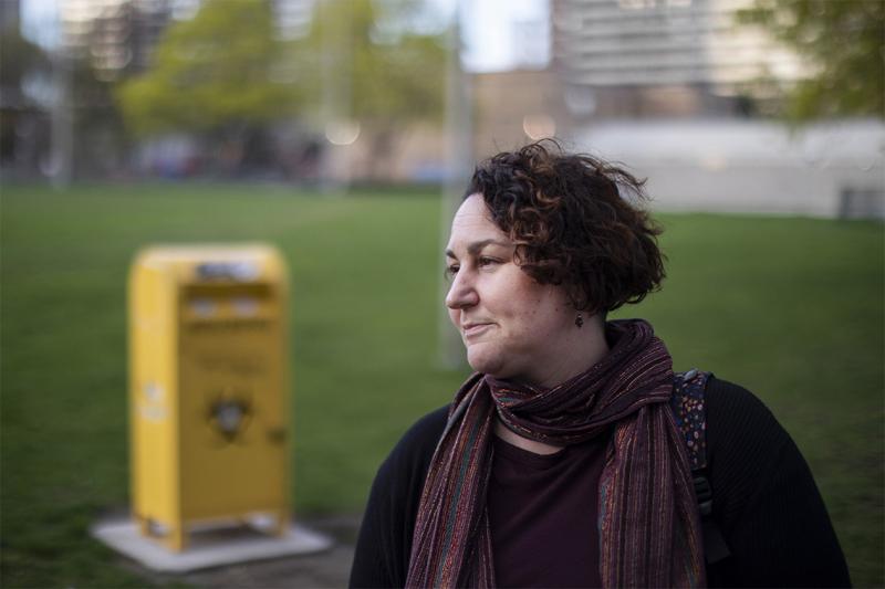 Gillian Kolla looks pensive, standing on the grass in Moss Park in front of a needle drop box.