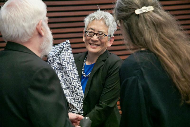 Carol Chin smiles, holding a wrapped present, as two people congratulate her.
