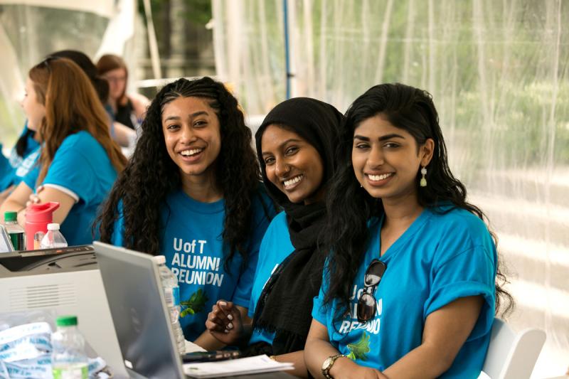 Three women wearing Alumni Reunion T-shirts smile from behind a registration desk.