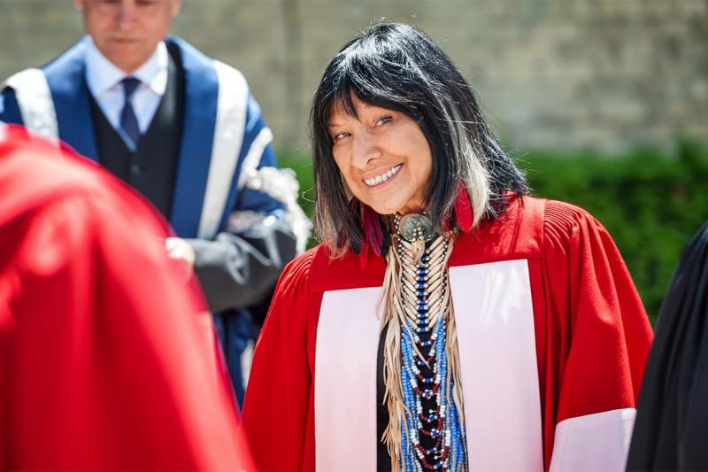 Buffy Sainte-Marie smiles mischievously as she looks through a crowd of dons, wearing academic robes.