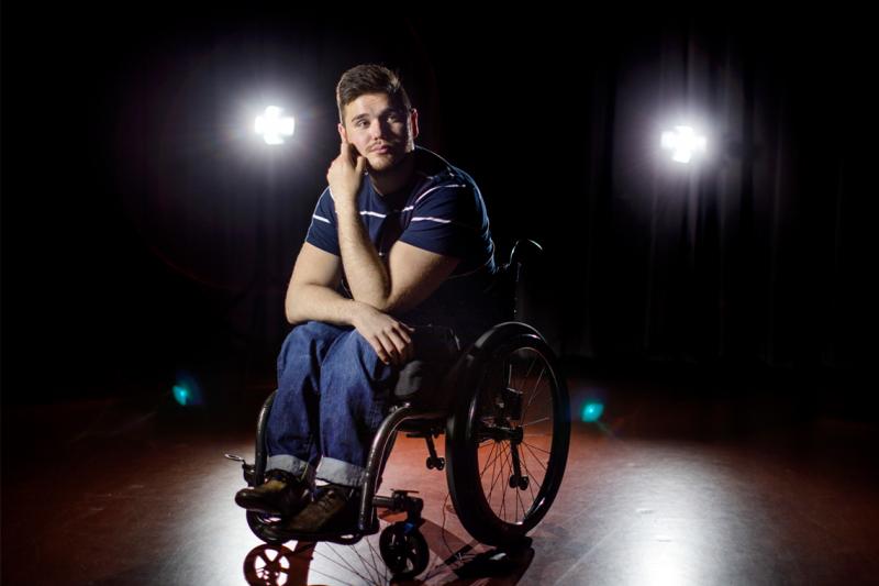 George Alevizos sits in his wheelchair, looking thoughtful, in the spotlight on a bare stage.