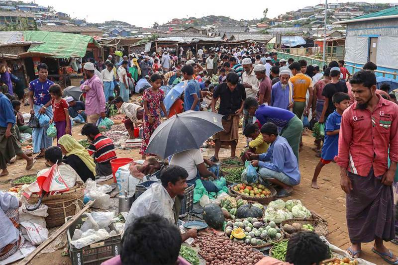People walk through a crowded, open-air market, between roughly-built shacks, as vendors display food in baskets and sacks.