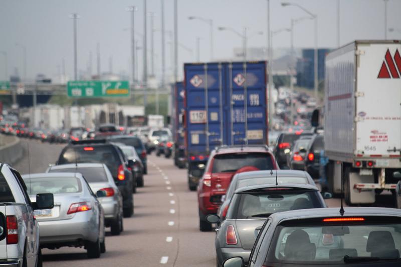 Cars sit in bumper-to-bumper traffic behind transport trucks on a highway in Toronto, before a hazy sky (photo by Danielle Scott via Flickr)