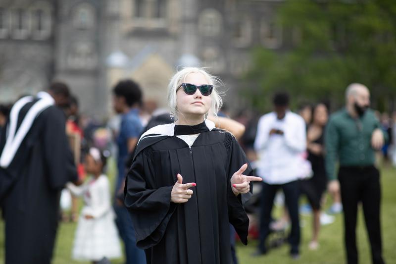 Elspeth Arbow makes finger guns as she stands on Front Campus in her academic robes and large sunglasses.