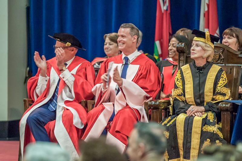 Mark Tewksbury grins and applauds while sitting on stage at Convocation Hall with Rose Patten, wearing academic robes.