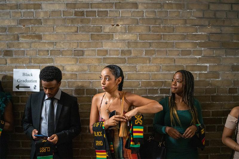 Graduates line up next to a sign that reads: Black Graduation. They carry their stoles over their arms.