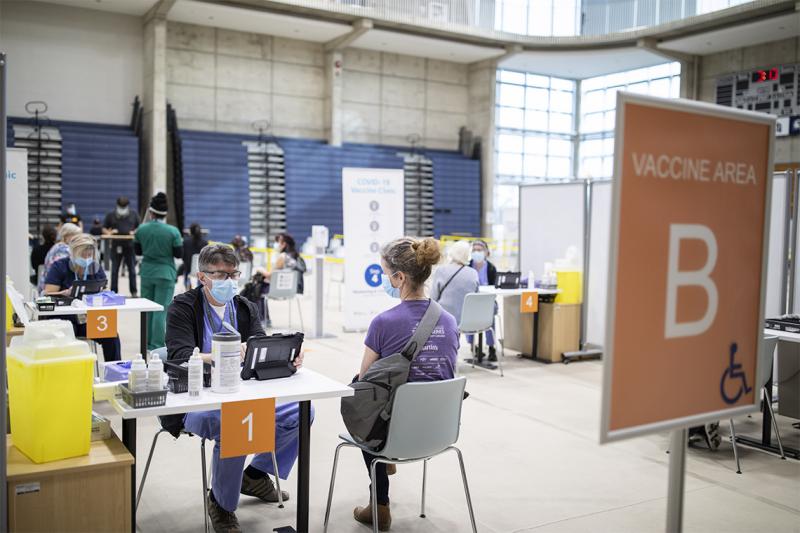 A masked patient and a masked volunteer sit at a table under a sign reading Vaccine Area B.