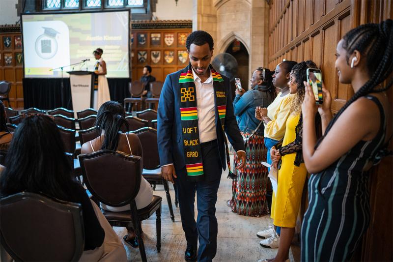 The audience claps and takes pictures as Isaiah Kidane walks down an aisle, wearing his Black Grad stole.