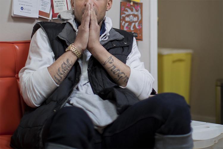 Cuz is shown from mouth to knees - he wears a vest and hoodie and holds his hands, in prayer position, in front of his face.