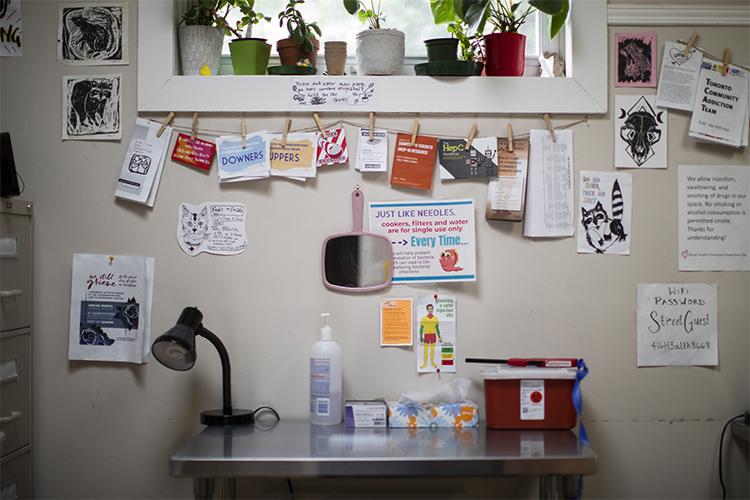 A small scrubbed table holds tissues, hand sanitizer and a box of medical supplies, beneath a window decorated with plants, art and info leaflets.