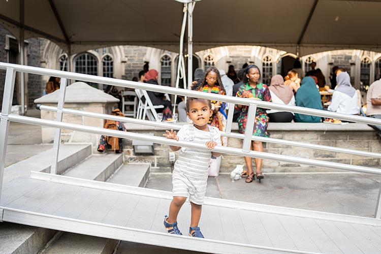 A toddler, Musa Hersi, peers through the railings of an accessibility ramp while adults chat at tables in the background.