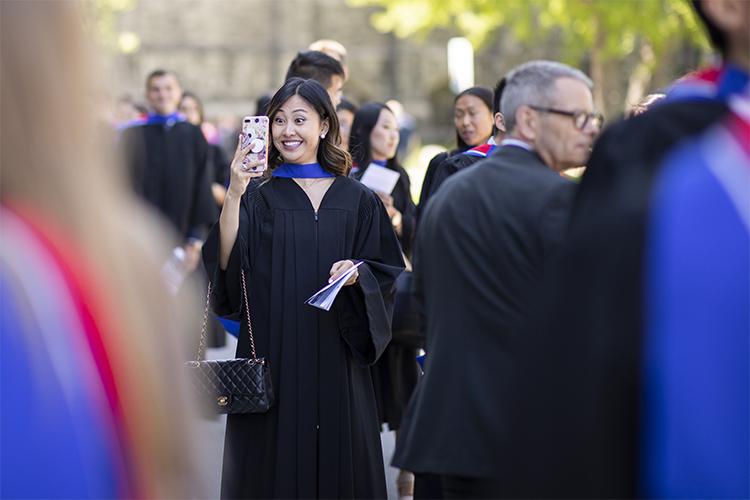 A woman wearing academic robes makes an excited, wide-eyed face as she takes a selfie.