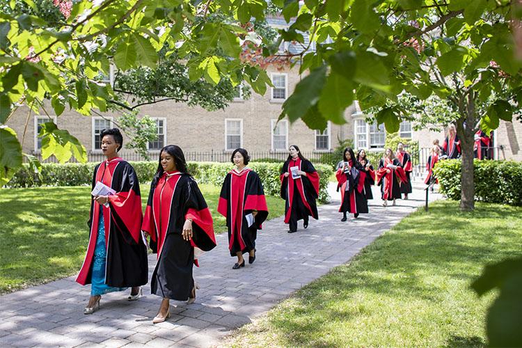 A line of students in academic robes walk across a sunlight garden.