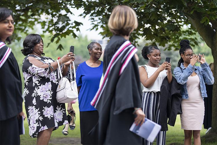 Smiling women hold up their phones as students in academic robes walk by.