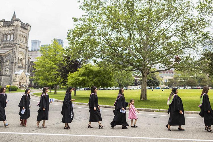 In the middle of a line of graduates in academic robes, a little girl walks holding one student's hand.
