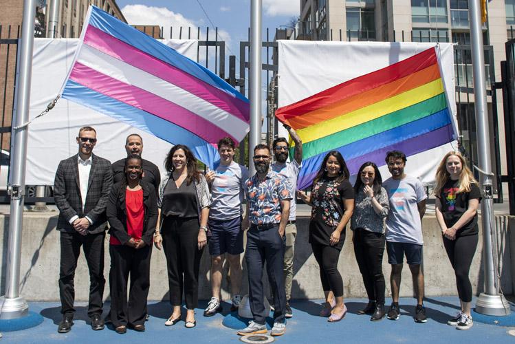 A group of people smile, standing at the base of two flagpoles with the Pride and Trans flags attached, ready for hoisting.