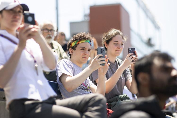 Three people sitting in a crowd on bleachers hold up their phones.