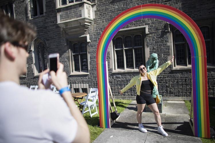 A man with a cellphone snaps a woman throwing out her arms as she stands in an archway painted to look like a rainbow.