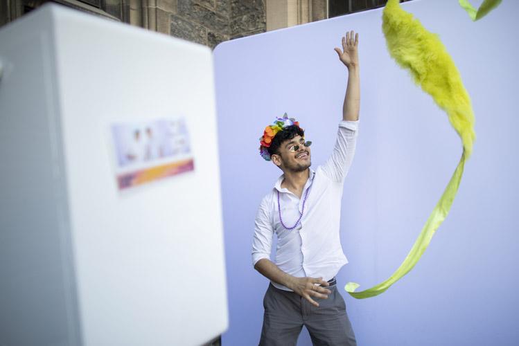 A man in a flower crown tosses a brightly-coloured streamer in the air while standing in a photo booth.