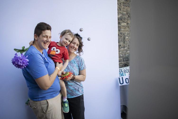 Two moms smile and laugh, holding their small child, who is wearing an Elmo T-shirt and holding a bright plastic flower.