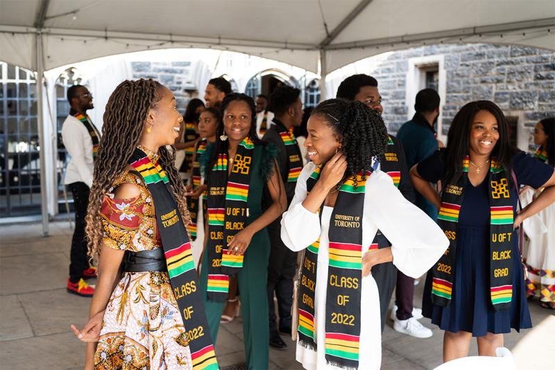 Graduates chat and laugh under a tent. Everyone is wearing a stole with the text: U of T Black Grad Class of 2022.