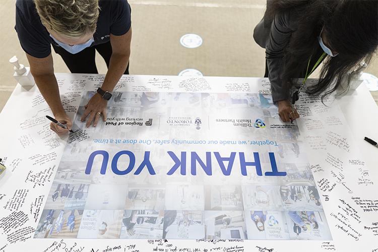 Two women write messages on a large poster. In the centre of the poster are the words: Thank you.