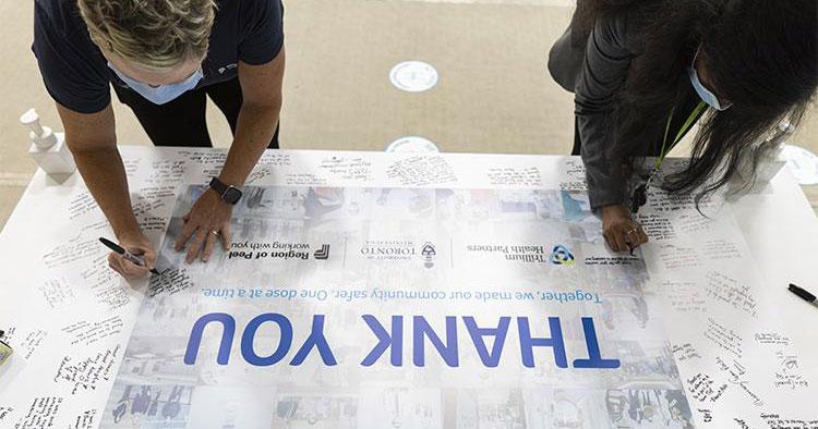 Two women write messages on a large poster. In the centre of the poster are the words: Thank you.