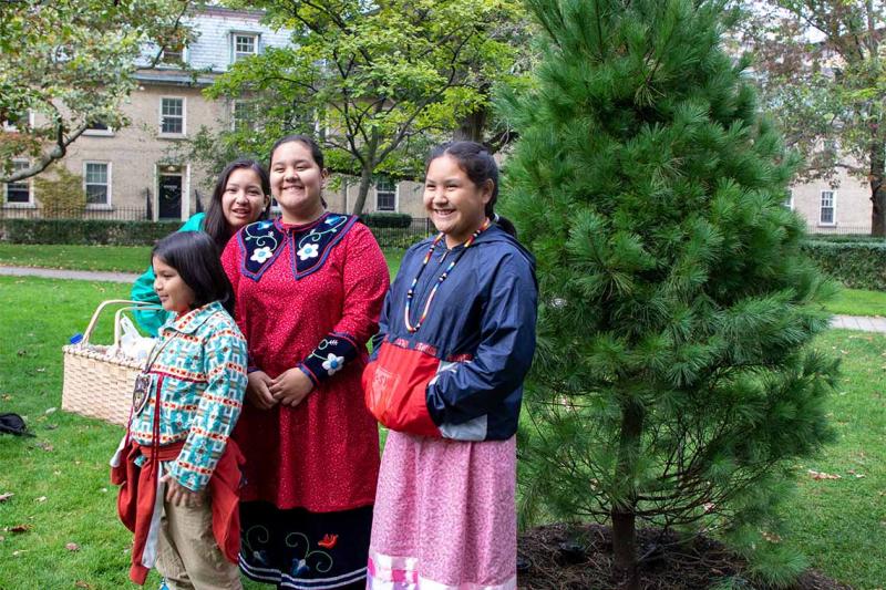 Four people in traditional Indigenous clothes smile while standing by a newly planted Eastern white pine tree.