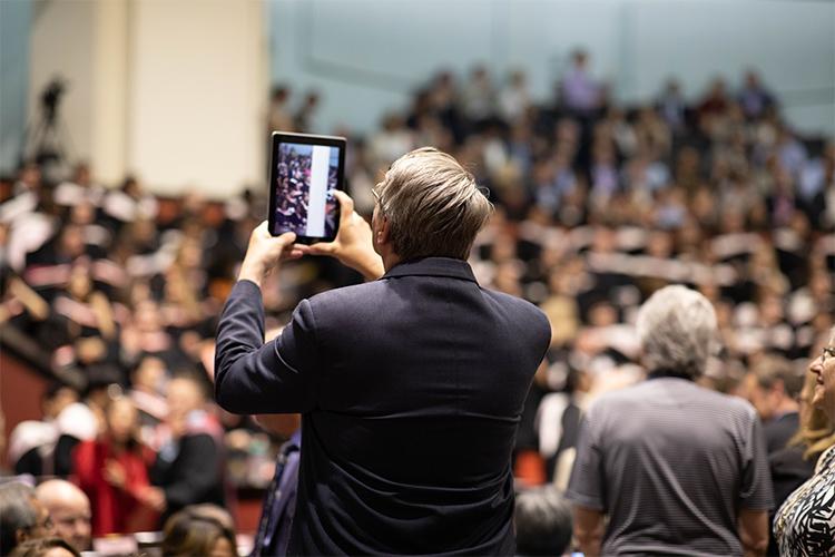 In Convocation Hall, a man holds up an tablet with the audience showing on screen.