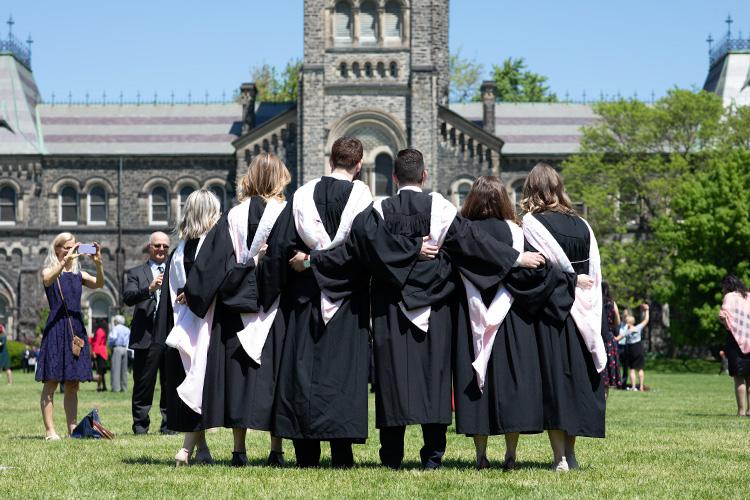 Seen from the back, six graduates in robes and gowns put their arms around each other for a picture.