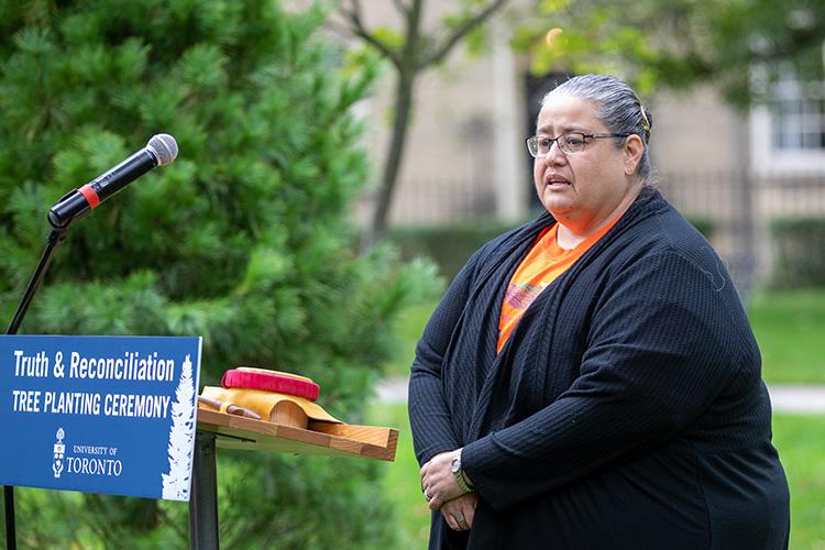 Susan Hill stands in front of a podium with a sign reading: Truth and Reconciliation Tree Planting Ceremony