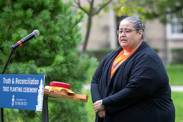 Susan Hill stands in front of a podium with a sign reading: Truth and Reconciliation Tree Planting Ceremony