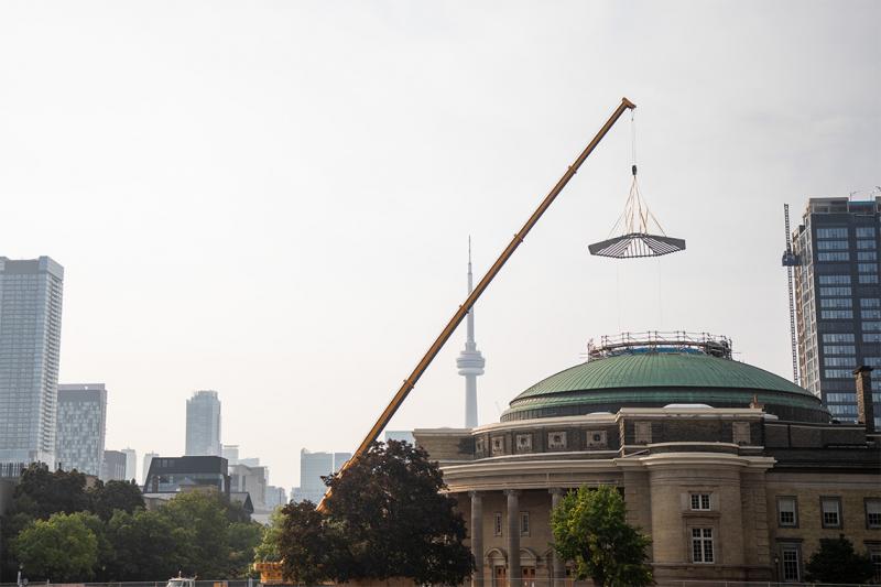 A circular glass skylight dangles from a crane over the roof of Convocation Hall.