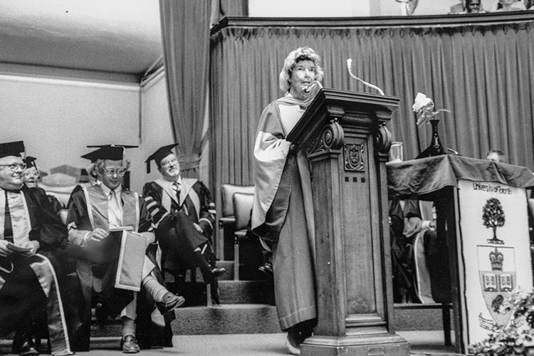 June Callwood, wearing academic robes, speaks from a lectern on stage with a row of dignitaries behind her.