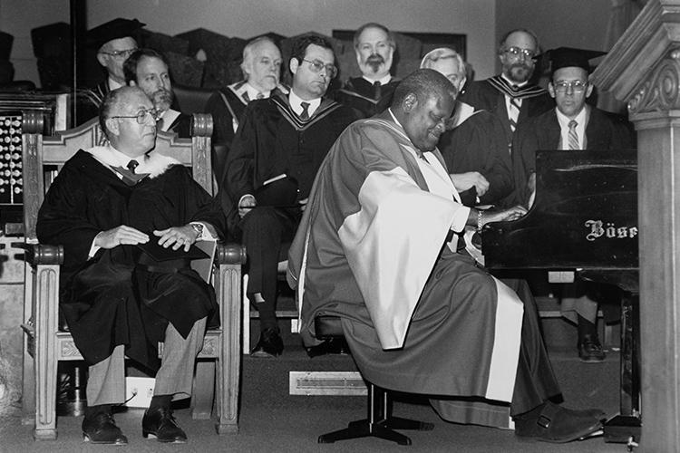 Oscar Peterson smiles as he plays piano on stage at Convocation Hall with the dignitaries behind him watching raptly.