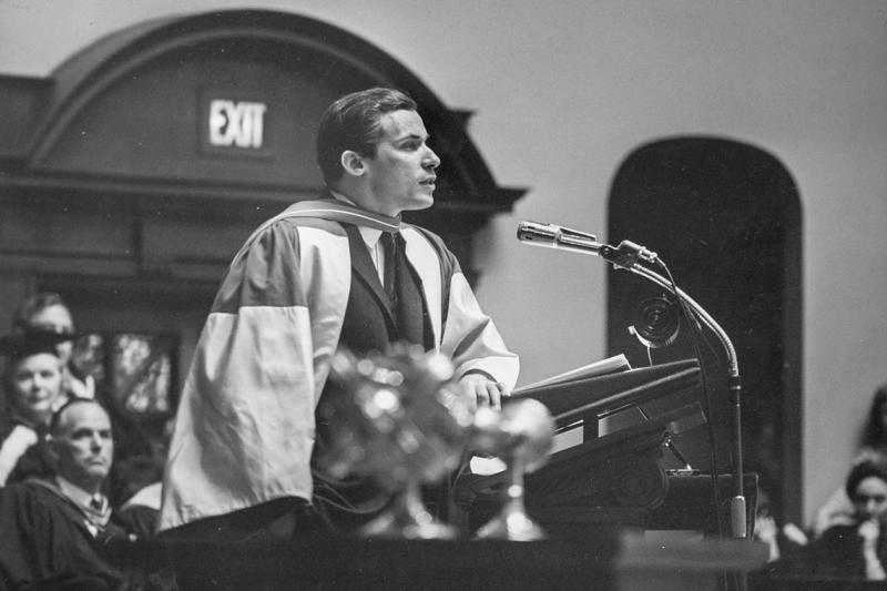 Glenn Gould, wearing academic robes, speaks at a lectern in Convocation Hall.
