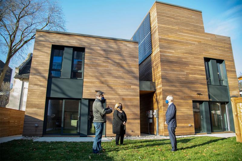 Meric Gertler, Scott Mabury and Gloria Cuneo wear masks as they stand apart on grass beside two wood-panelled houses.