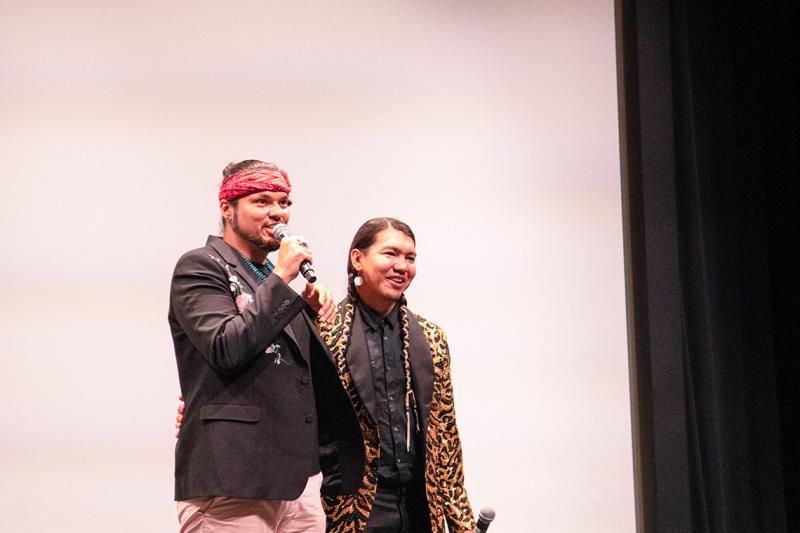 Anthony Johnson and James Makokis stand with arms around each other while speaking into a microphone on stage.