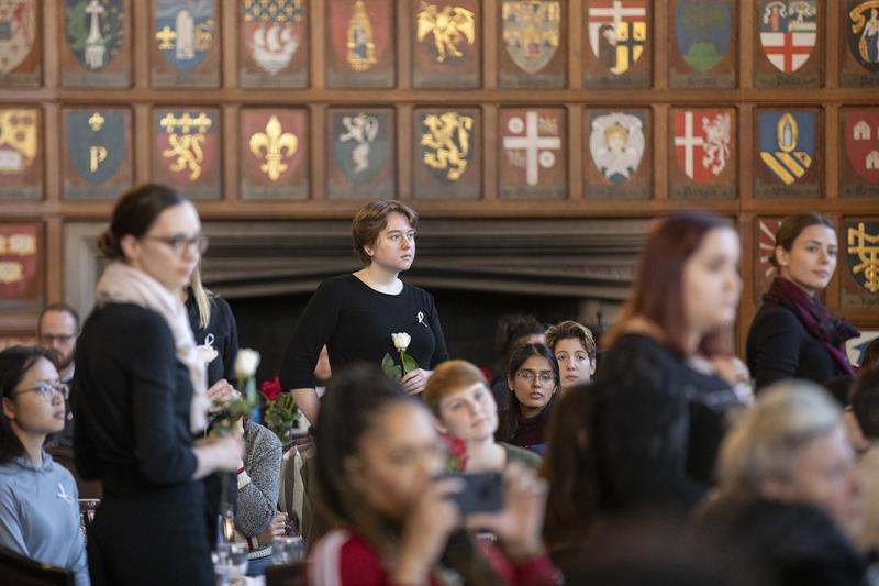 Women dressed in black and holding roses listen attentively, in a room in Hart House.