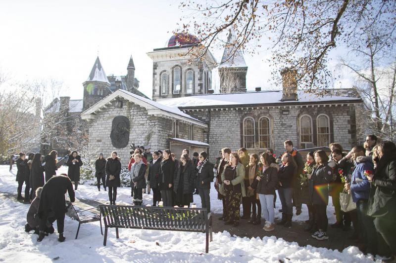 A crowd stands in the snow east of University College, watching solemnly as women place fresh roses on two metal benches.