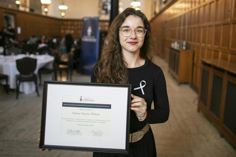 Sydney Narciso Wilson holds up a framed certificate, standing in a room in Hart House.