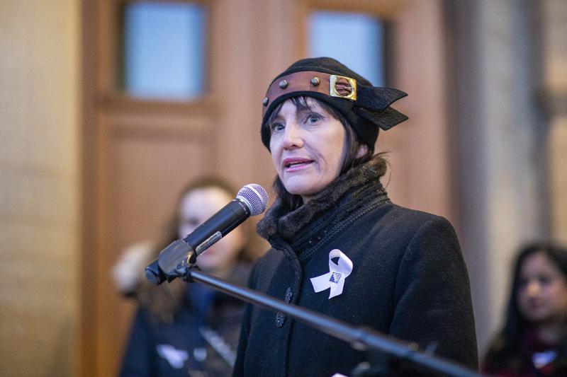 Marisa Sterling looks serious as she speaks into a microphone, wearing a white ribbon.