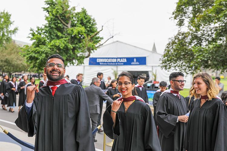 A smiling man and woman in academic robes extend their pinky fingers, which are wearing plain iron rings.