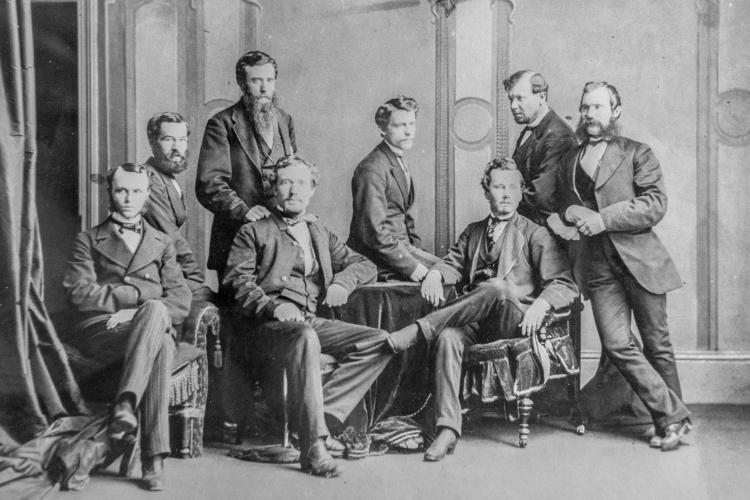 Eight men in three-piece suits and 1870s hairstyles lounge around a cloth-draped table.