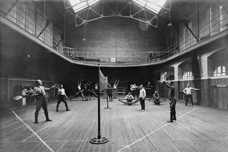 Four men play badminton in a gym while men behind them work out on rowing machines and other equipment.