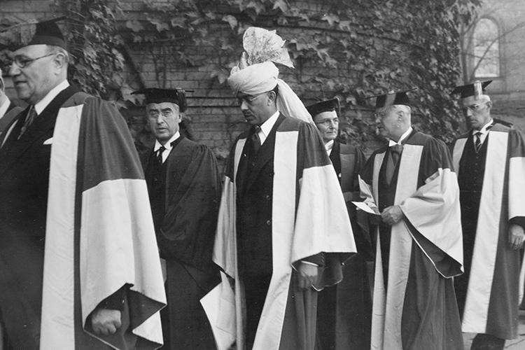A man wearing a turban with large cockade walks in a procession with several older men in academic robes.
