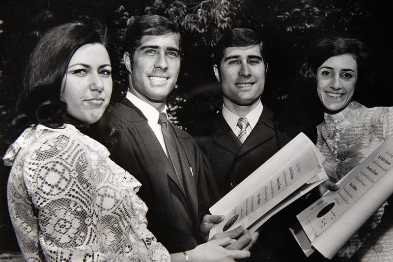 Edward and Jay Keystone, in academic robes, smile and hold their graduation certificates while flanked by two women.