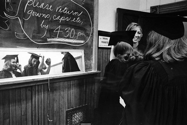 A mirror propped on a blackboard reflects two smiling women, one of whom holds her hand to her mortarboard hat.