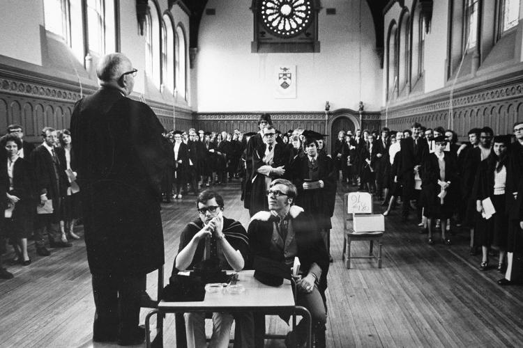 Students in academic robes stand around the edges of a high-ceilinged room and watch a man who is calling out instructions.